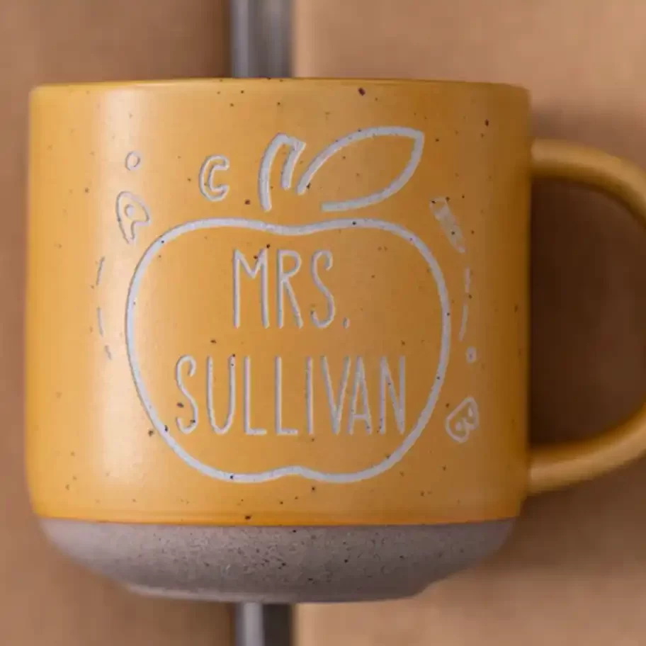 A ceramic mug with the teacher's name' text, decorated with colorful apples and pencils. Perfect as a personalized gift, it rests on a wooden surface.