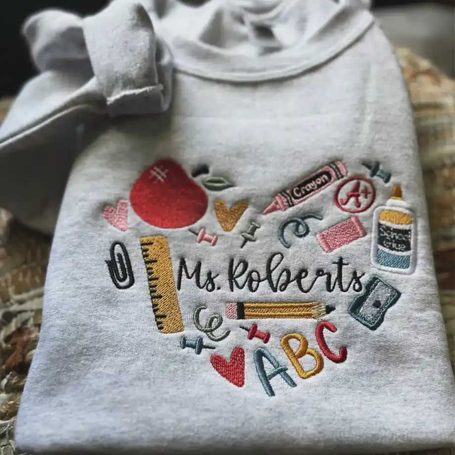 A cozy sweatshirt with the text 'Best Teacher Ever' and playful designs like books and apples. Perfect for gifting, it’s displayed neatly on a wooden surface.