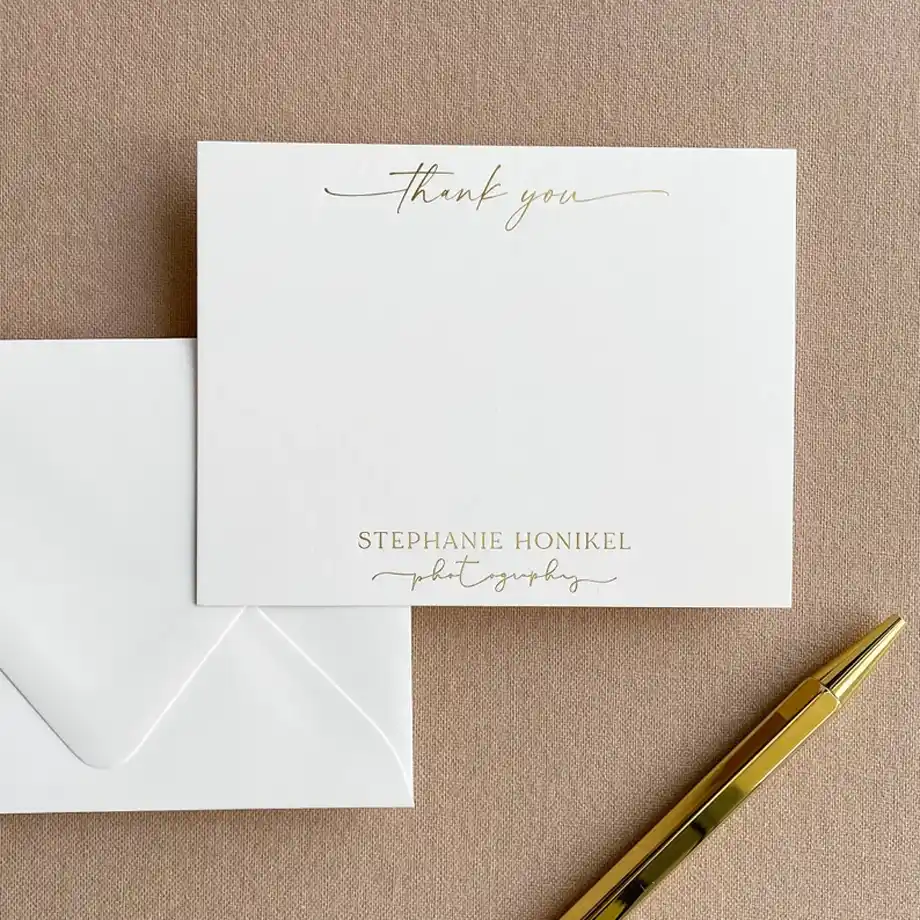 A minimalist thank you card with gold lettering, featuring the text "Thank You" and personalized branding for a photographer. The card is placed on a neutral beige background with a matching envelope and gold pen, perfect for writing a thoughtful thank you message for a gift.