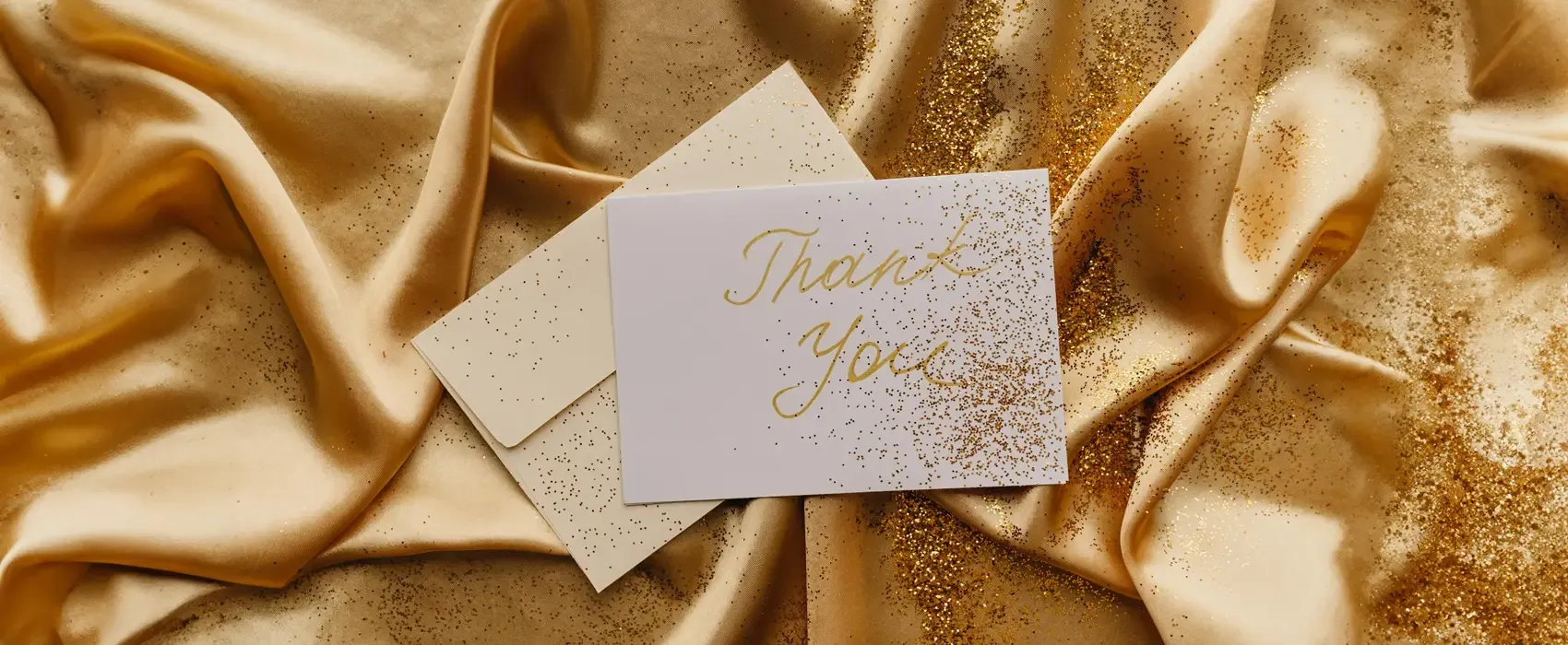 A luxurious thank you card with "Thank You" written in gold script, set on shimmering gold satin fabric sprinkled with glitter, perfect for expressing a heartfelt thank you message for a gift.