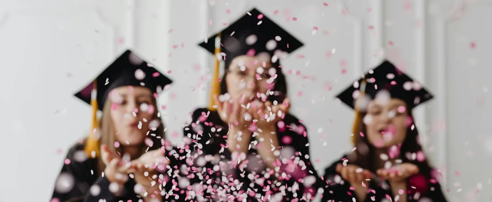 Three joyful graduates in black caps and gowns celebrating their achievement by blowing pink and white confetti, symbolizing success and inspiration for graduation quotes.