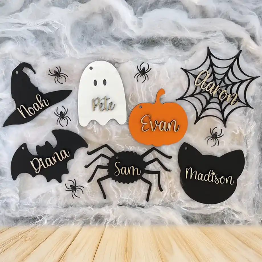 A collection of custom Halloween name tags in spooky shapes like a witch hat, ghost, pumpkin, spider, bat, and cat. Each tag features a name in stylish script, perfect for decorating a boo basket. The tags are displayed on a cobweb-covered background with small black spider accents.