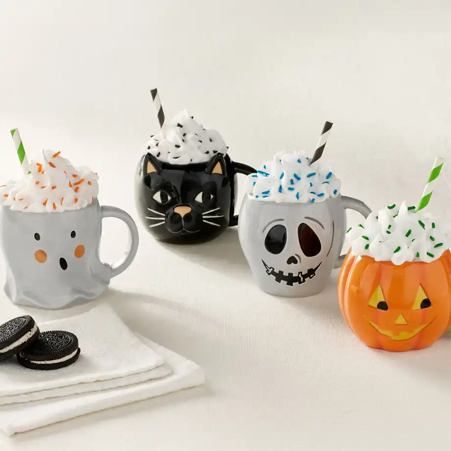 A festive set of Halloween-themed mugs featuring a ghost, black cat, skull, and pumpkin, each topped with whipped cream and sprinkles—an adorable and cozy addition to any Halloween boo basket. 