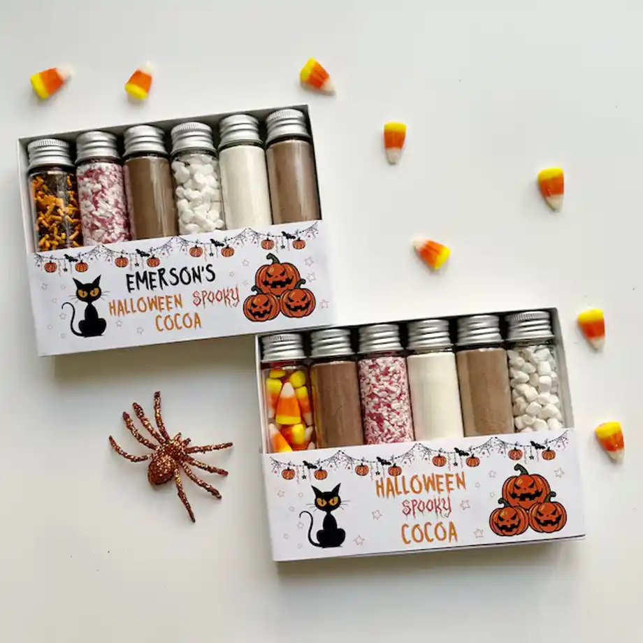 Two festive Halloween cocoa kits with jars of hot chocolate mix, candy corn, marshmallows, and sprinkles, decorated with pumpkins and black cats—perfect for warming up your Halloween boo basket.