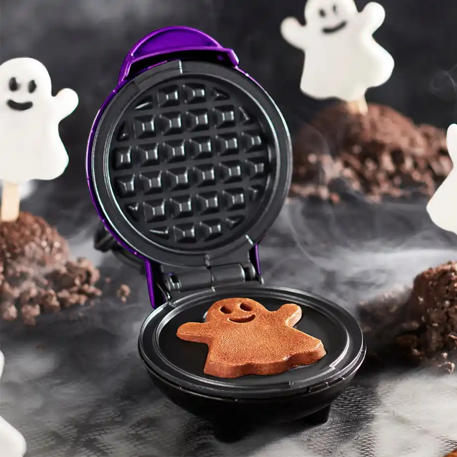 A festive mini waffle maker creating a cute, ghost-shaped waffle, surrounded by Halloween-themed decorations—perfect for adding a spooky and sweet touch to any Halloween boo basket.