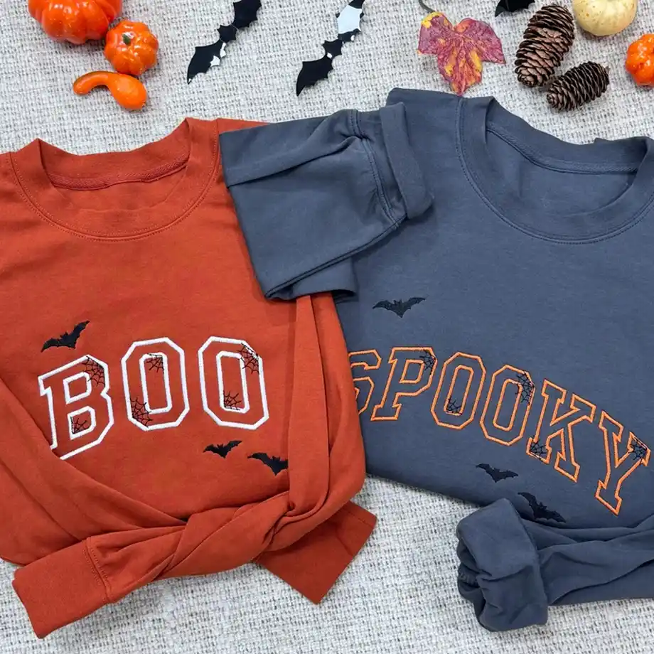 Two Halloween-themed shirts laid flat, one orange with "BOO" and one gray with "SPOOKY," both decorated with bats and cobweb designs — festive, cozy, and perfect for adding a stylish touch to any Halloween boo basket.