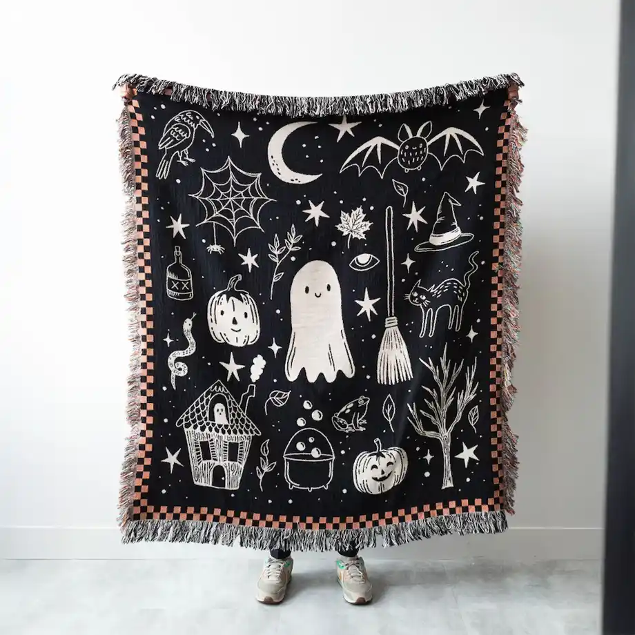 A whimsical Halloween throw blanket featuring adorable ghosts, moons, bats, and a checkered orange and black design—perfect for snuggling up on a chilly night or adding a cozy touch to a Halloween boo basket.