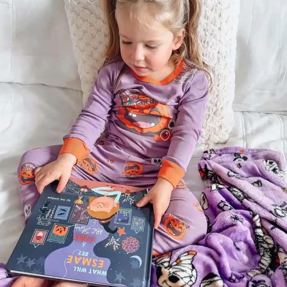 A young girl in festive Halloween pajamas sits on a white bed, holding a personalized Halloween-themed book. She is wrapped in a cozy purple mummy-print blanket, creating the perfect spooky-yet-snuggly vibe. This adorable moment showcases a thoughtful boo basket idea, perfect for making Halloween magical with books, pajamas, and cozy treats.