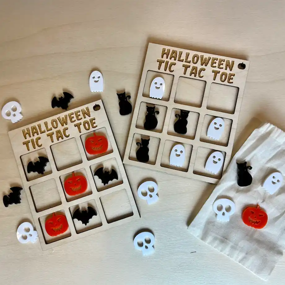 Two wooden Halloween-themed tic-tac-toe boards with spooky game pieces, including ghosts, pumpkins, bats, black cats, and skulls, are displayed on a light wooden surface. A small fabric bag holds extra game pieces, making this a fun and festive addition to any boo basket for kids or adults.