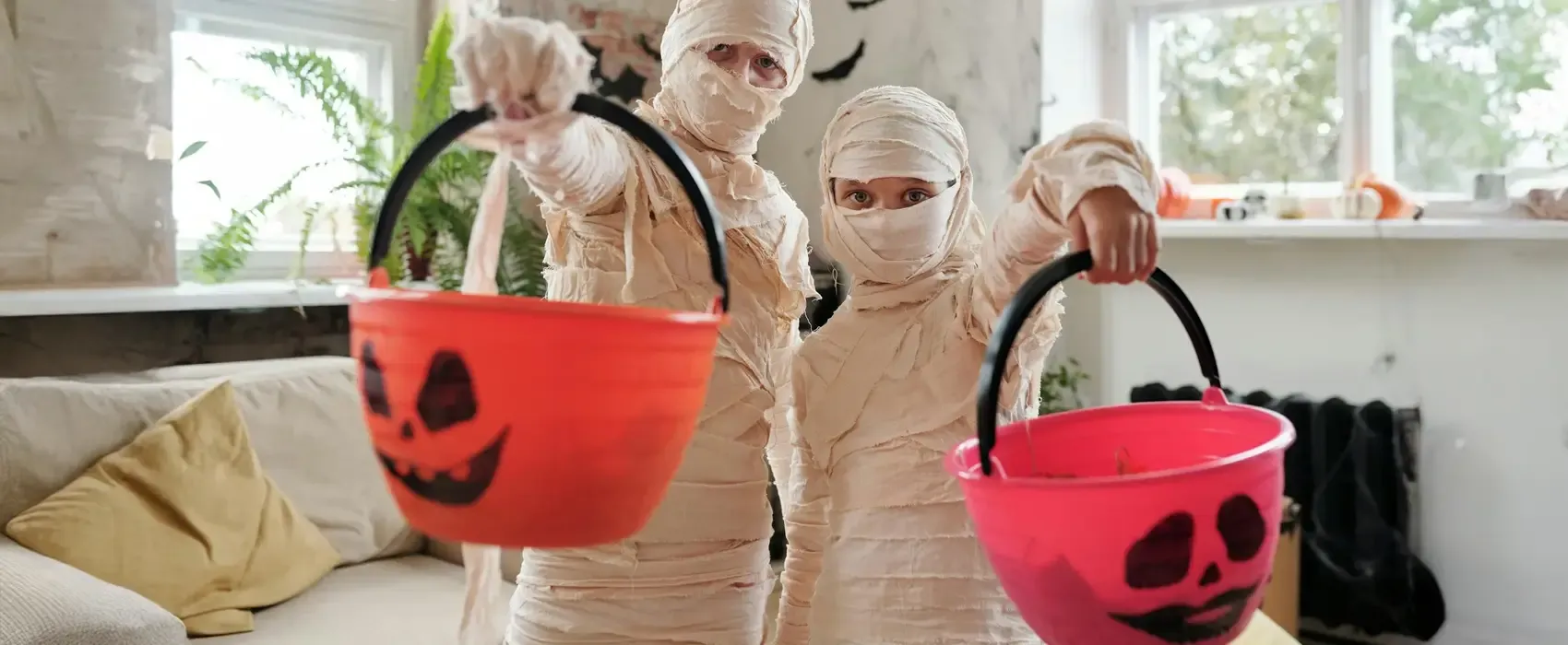 Two children dressed as mummies hold out Halloween-themed boo baskets with jack-o'-lantern faces, ready for trick-or-treating in a festively decorated home.