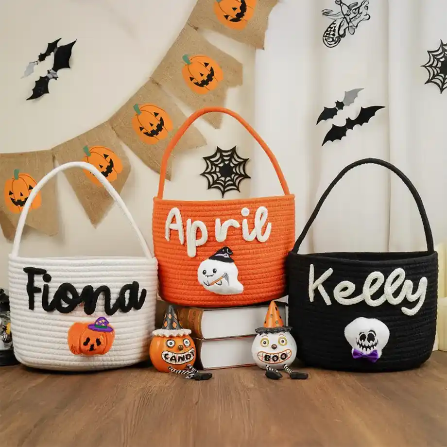 Three personalized rope boo baskets in white, orange, and black, featuring embroidered names and Halloween-themed decorations like pumpkins, ghosts, and skulls. The baskets are displayed with festive Halloween decor, including a pumpkin banner, bat cutouts, and spooky figurines.