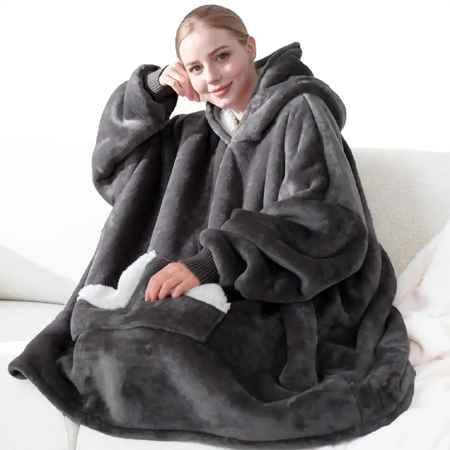 A woman relaxing on a couch wearing a plush, oversized hoodie blanket in dark gray with a fluffy sherpa lining. This cozy and warm wearable blanket makes for one of the best white elephant gift ideas, perfect for winter lounging and comfort.