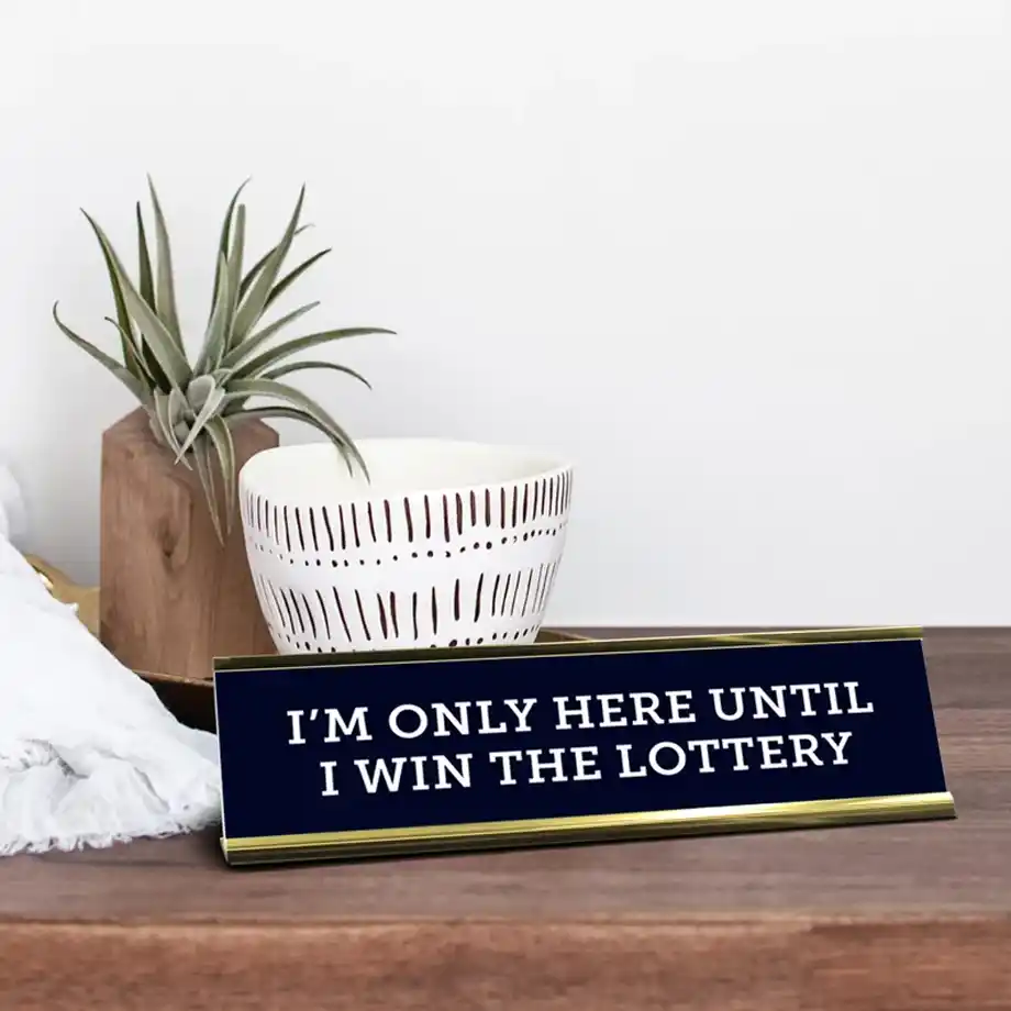 A navy blue and gold desk nameplate with the humorous message "I’M ONLY HERE UNTIL I WIN THE LOTTERY." Placed on a wooden desk with modern decor, this novelty office sign makes a perfect choice for white elephant gift ideas, bringing laughter to any workplace or gift exchange.