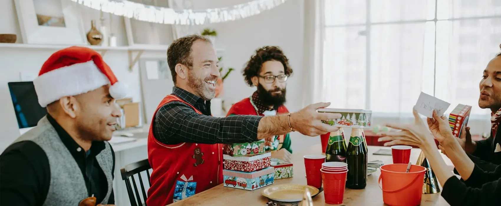 A group of friends at a festive holiday party exchanging presents, laughing, and enjoying a white elephant gift exchange with Christmas-themed wrapping and drinks on the table.