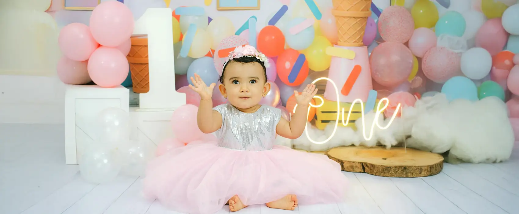 A baby girl in a silver and pink tutu dress sits in front of a colorful ice cream-themed birthday setup with balloons, an ice cream cone backdrop, and a glowing "One" sign. A sweet inspiration for first birthday themes for girls.