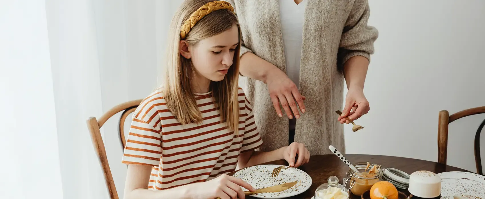 A young girl in a striped shirt sits at a dining table, looking down at her nearly empty plate with a serious expression. A woman, possibly her mother, stands beside her, holding a small spoon and gesturing toward the plate. The table has a few jars of peanut butter, oranges, and other food items. The scene captures the restrictive food environment often associated with almond mom tendencies.