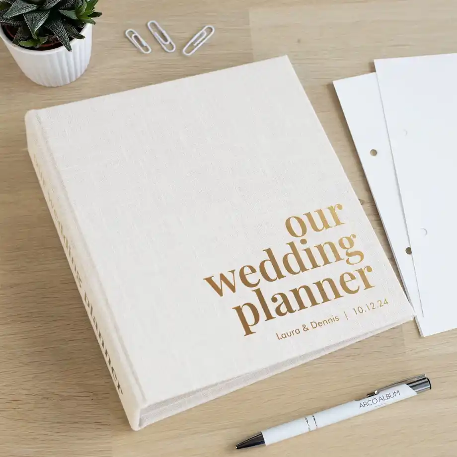 A stylish wedding planner with gold lettering on a wooden desk, surrounded by a pen and paper clips. A thoughtful choice for engagement gift ideas to help couples plan their big day.