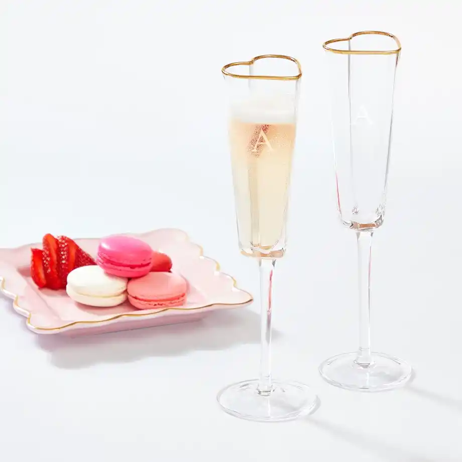 A set of two heart-shaped champagne flutes with gold rims and engraved initials, perfect for celebrating special moments. Paired with a pink plate of colorful macarons and fresh strawberries, this elegant glassware makes a stylish and romantic choice for engagement gift ideas.