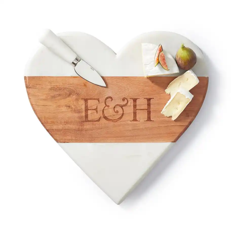 A heart-shaped cheese board made of marble and wood, engraved with initials for a personalized touch. Accompanied by a cheese knife, brie, and fresh figs, this elegant and practical piece makes a perfect choice for engagement gift ideas.