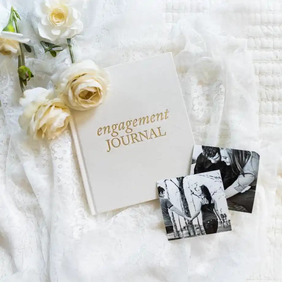 A beautifully designed engagement journal with gold lettering, placed on delicate lace fabric alongside white roses and black-and-white couple photos. A sentimental and stylish choice for engagement gift ideas.