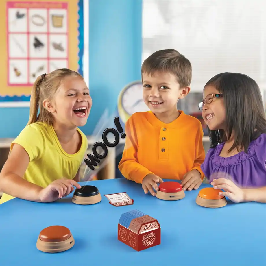 Three smiling children are playing a fun Indoor Games activity involving sound buzzers and a card-based challenge. The game encourages laughter and interaction as one child presses a button that makes a "Moo!" sound. This engaging and educational Indoor Games option is perfect for kids' playtime, helping with learning and social skills while having fun indoors.