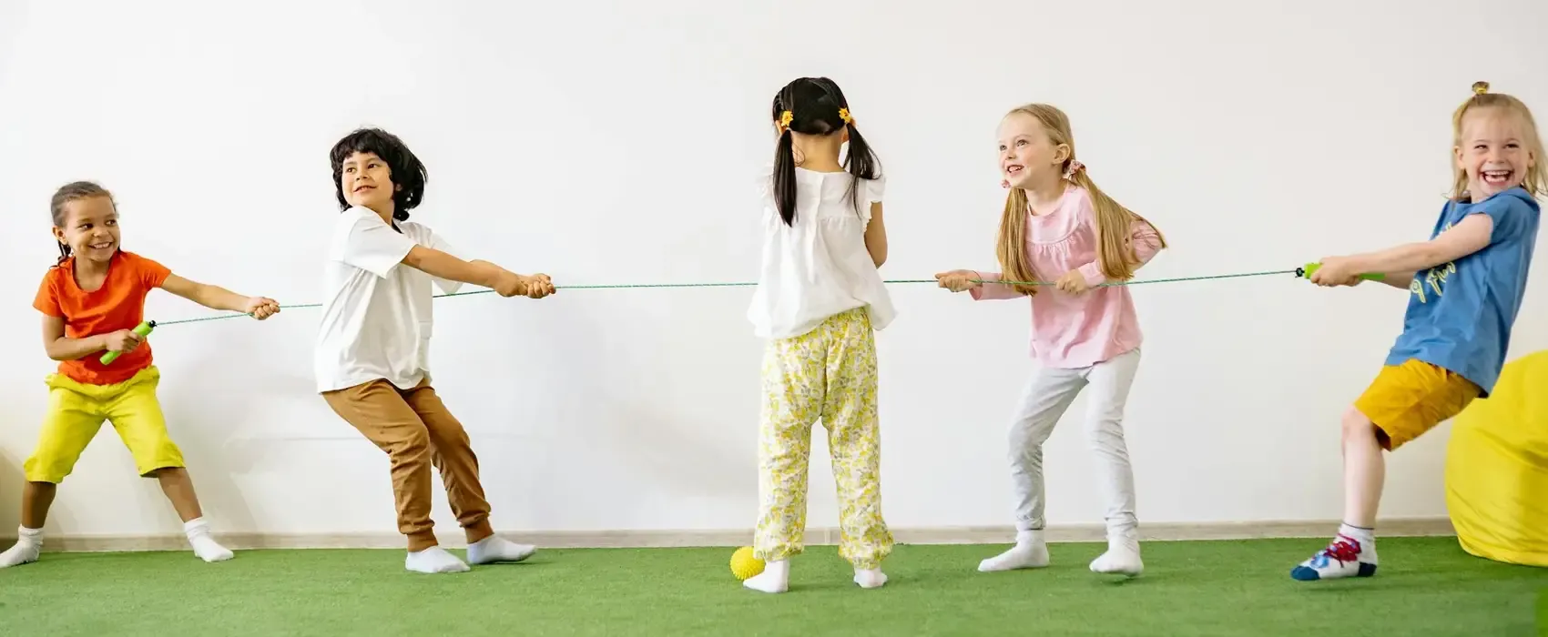 A group of five happy children playing a tug-of-war game indoors on a green carpet. They are pulling a rope with excitement and energy, showcasing a fun and engaging activity. This image highlights the joy of Indoor Games for kids.