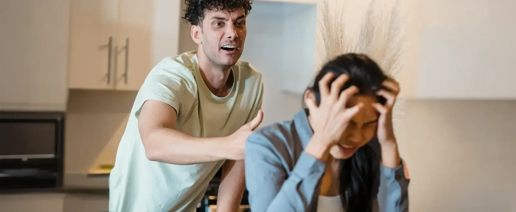 A distressed woman sits in a kitchen holding her head in frustration while a man angrily gestures at her illustrating red flags in a relationship such as emotional abuse and poor communication