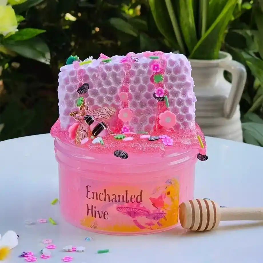 A colorful pink slime labeled “Enchanted Hive,” decorated with glitter, flower sprinkles, and a bee charm, sitting on a white surface with a honey dipper — perfect for visualizing how to get slime out of clothes after playtime fun.